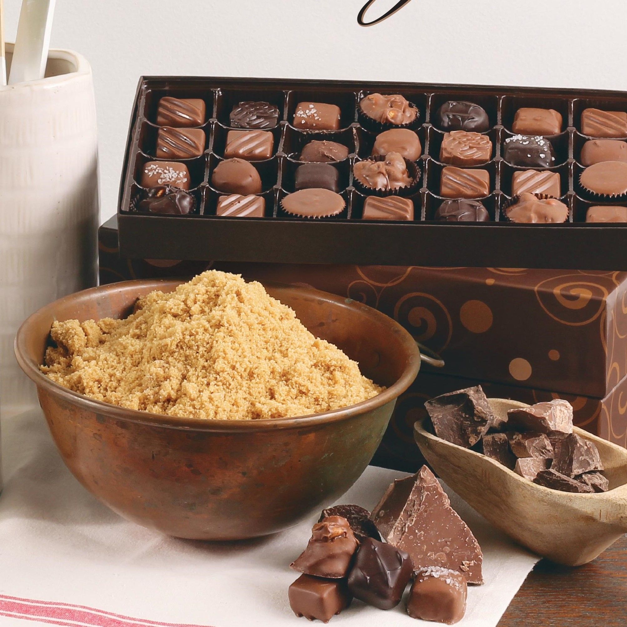 A box of assorted gourmet chocolates displayed with a rustic copper bowl filled with brown sugar, a small wooden dish of chocolate chunks, and various chocolate pieces on a white napkin.
