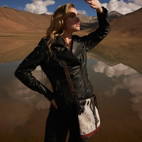 Woman in a studded black leather jacket posing in a desert landscape, shading her eyes while carrying a stylish cowhide crossbody bag.