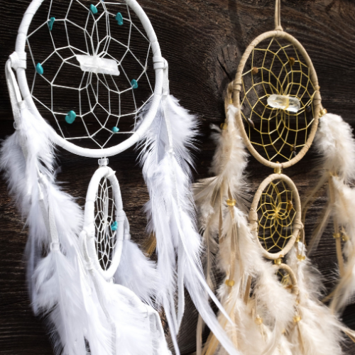 Two handmade dreamcatchers hanging against a wooden background, adorned with white and beige feathers, beads, and gemstone accents.