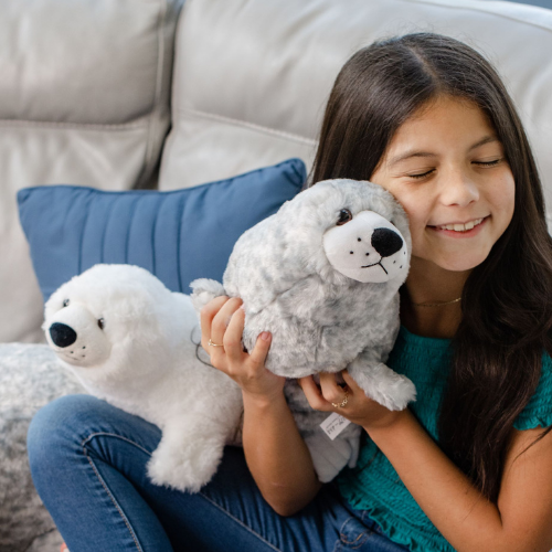 Young girl smiling with her eyes closed while hugging two soft plush toys—a gray seal and a white polar bear—on a cozy couch.