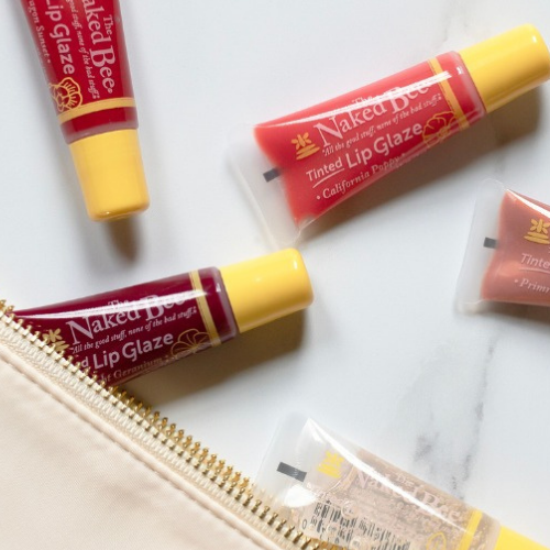 Flat lay of Naked Bee tinted lip glaze tubes in various shades, partially spilling out of a cosmetic bag on a marble surface.