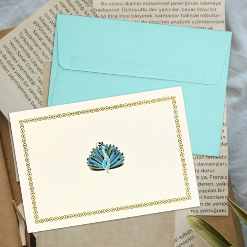 Elegant white notecard with a blue and teal peacock illustration and gold border, paired with a light blue envelope, displayed atop layered papers.