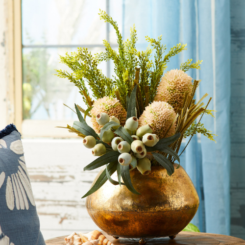 Shiny gold vase holding a realistic-looking arrangement of soft pink thistles, green pods, and foliage, displayed in a bright, coastal-themed room.
