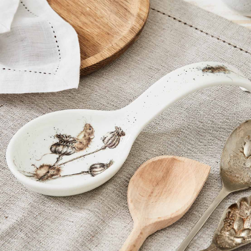 White ceramic spoon rest with painted botanical motifs of dried plants, displayed with wooden and metal utensils on a neutral linen tablecloth.