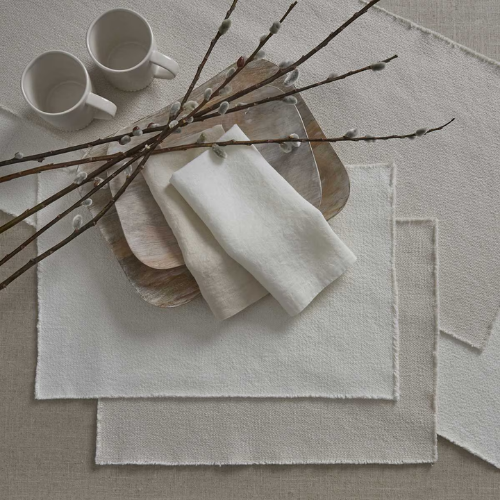 Elegant white textured placemats and napkins styled with neutral-toned dishware, ceramic mugs, and pussy willow branches on a light fabric background.