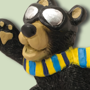 Playful bear figurine wearing aviator goggles and a yellow-and-blue striped scarf, raising its paw with a cheerful expression.
