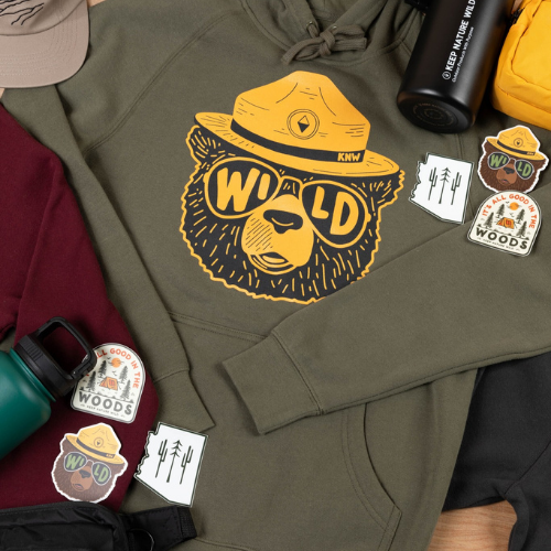 Olive green hoodie with a graphic of a bear wearing sunglasses and a ranger hat labeled “WILD,” surrounded by outdoor-themed stickers, water bottles, and gear.