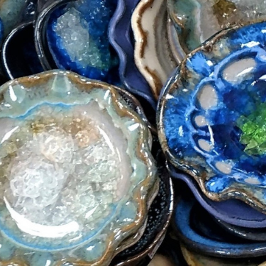 Collection of handcrafted ceramic dishes in various glazes, featuring rich blue, green, and crystal-like textures with organic, wavy edges.