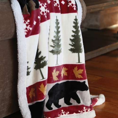 Soft fleece blanket draped over a couch, featuring a pattern of evergreen trees, snowflakes, maple leaves, and black bears on a red and white background.