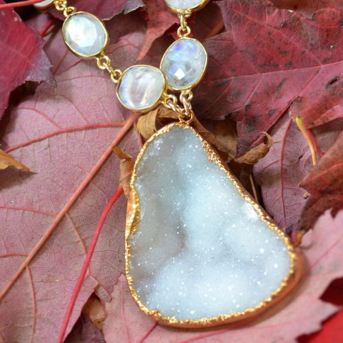 Close-up of a druzy crystal pendant edged in gold, paired with faceted gemstone links, laid on vibrant red autumn leaves.