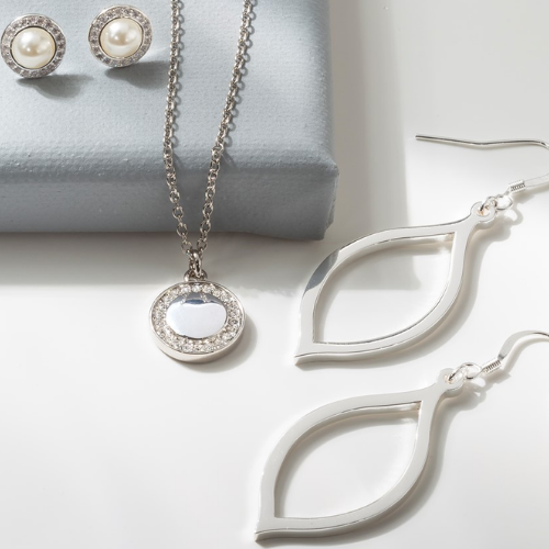 Close-up of elegant silver jewelry including pearl stud earrings, a pendant necklace with a crystal border, and open teardrop-shaped dangle earrings, displayed on a white surface.