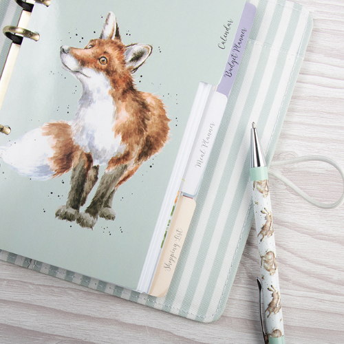 Open planner featuring a watercolor fox illustration, labeled dividers, and a matching fox-patterned pen on a woodgrain surface.