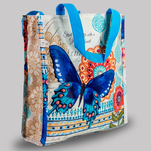 Reusable shopping tote bag with bright blue handles, decorated with a large blue butterfly and floral collage design in a mix of vibrant colors and handwritten-style text.