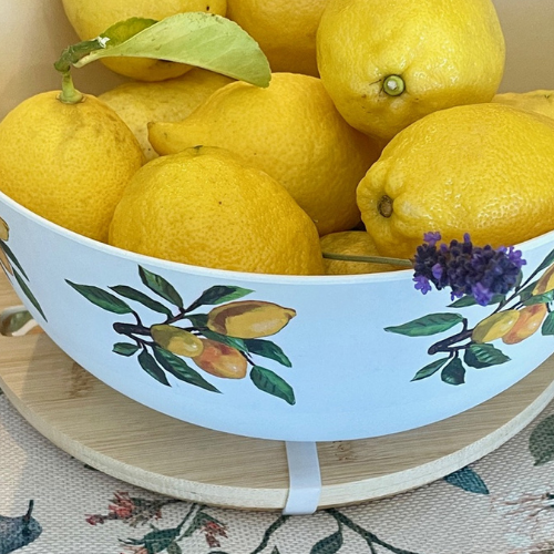 Fresh yellow lemons in a white bowl adorned with hand-painted lemon branch illustrations, placed on a wooden tray with a floral-patterned cloth beneath.