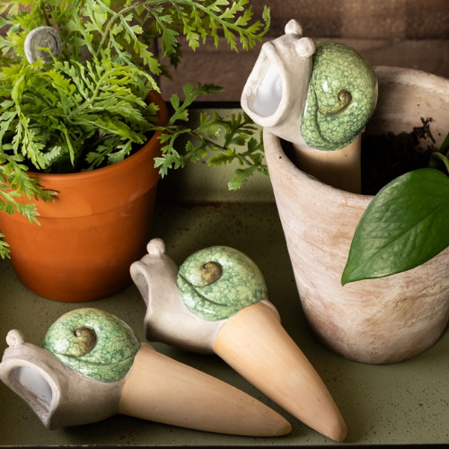 Set of whimsical ceramic snail-shaped watering stakes displayed beside potted plants, designed for slowly hydrating indoor greenery.