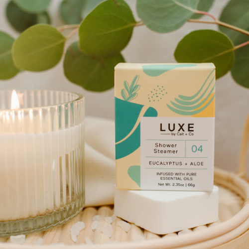LUXE brand eucalyptus and aloe shower steamer displayed on a wooden tray with a lit candle and fresh greenery in the background.