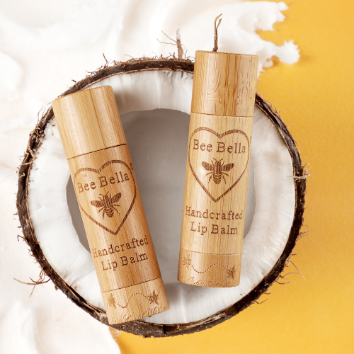 Two bamboo tubes of Bee Bella handcrafted lip balm with engraved logos, styled inside a halved coconut against a bright background with coconut shavings.