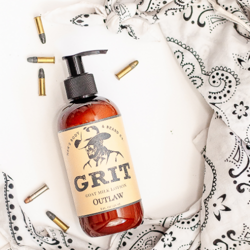 Amber bottle of Grit goat milk lotion by Outlaw, surrounded by a black-and-white bandana and scattered bullet casings for a rugged Western aesthetic.