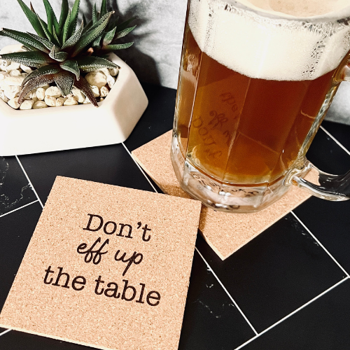 Close-up of a pint of beer resting on a cork coaster printed with the phrase “Don’t eff up the table,” next to a small succulent on a black tile surface.