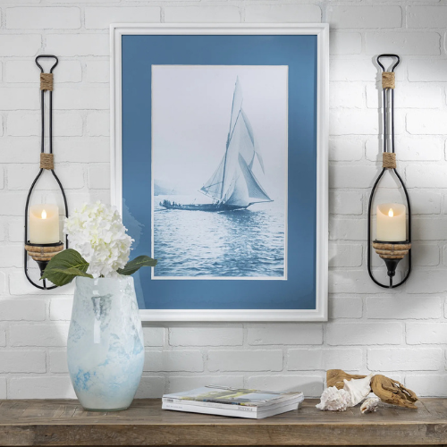 Coastal-themed interior display with a framed sailboat photograph in blue and white, flanked by two wall-mounted candle holders shaped like oars, and a blue and white vase with white hydrangeas on a rustic table.