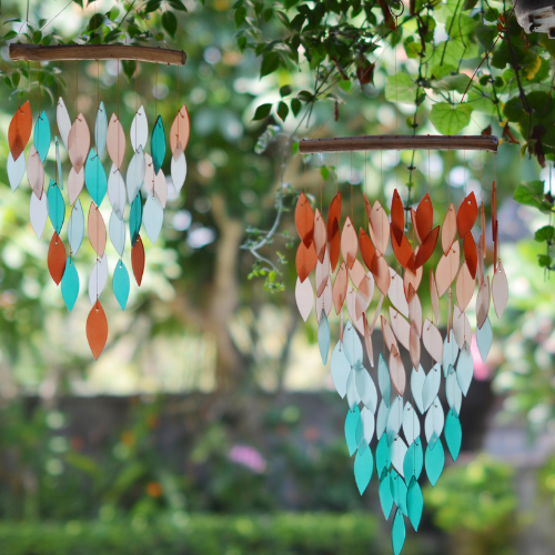 Two hanging wind chimes made of cascading leaf-shaped pieces in shades of teal, orange, and white, suspended from curved wooden bars in a lush garden setting.