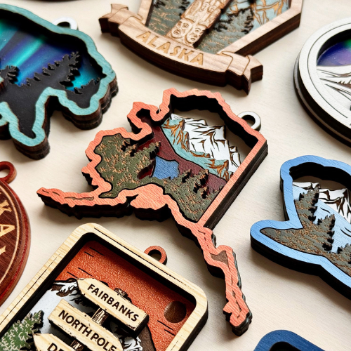 Detailed wooden ornaments shaped like the state of Alaska, decorated with mountain, forest, and northern lights scenes, including cutout tags labeled with destinations like “Fairbanks” and “North Pole.”