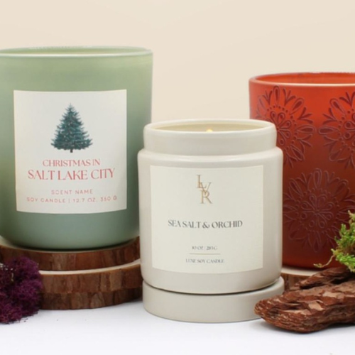 Trio of decorative soy candles in jars labeled with scents like “Christmas in Salt Lake City” and “Sea Salt & Orchid,” arranged with wood slices and natural elements like bark and moss.