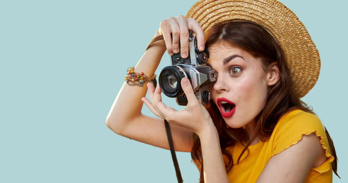 Surprised young woman in straw hat and yellow top holding vintage camera against light blue background.