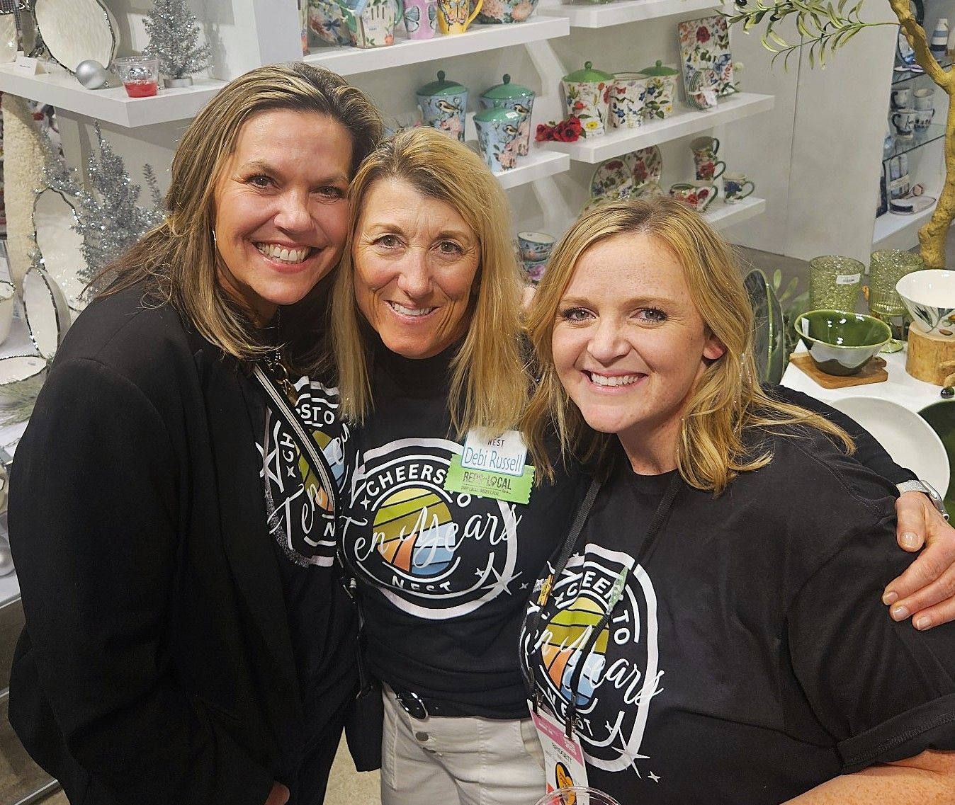 Three women posing together in a retail showroom filled with colorful ceramics and seasonal decor. They are all smiling and wearing matching “Cheers to Ten Years” shirts.