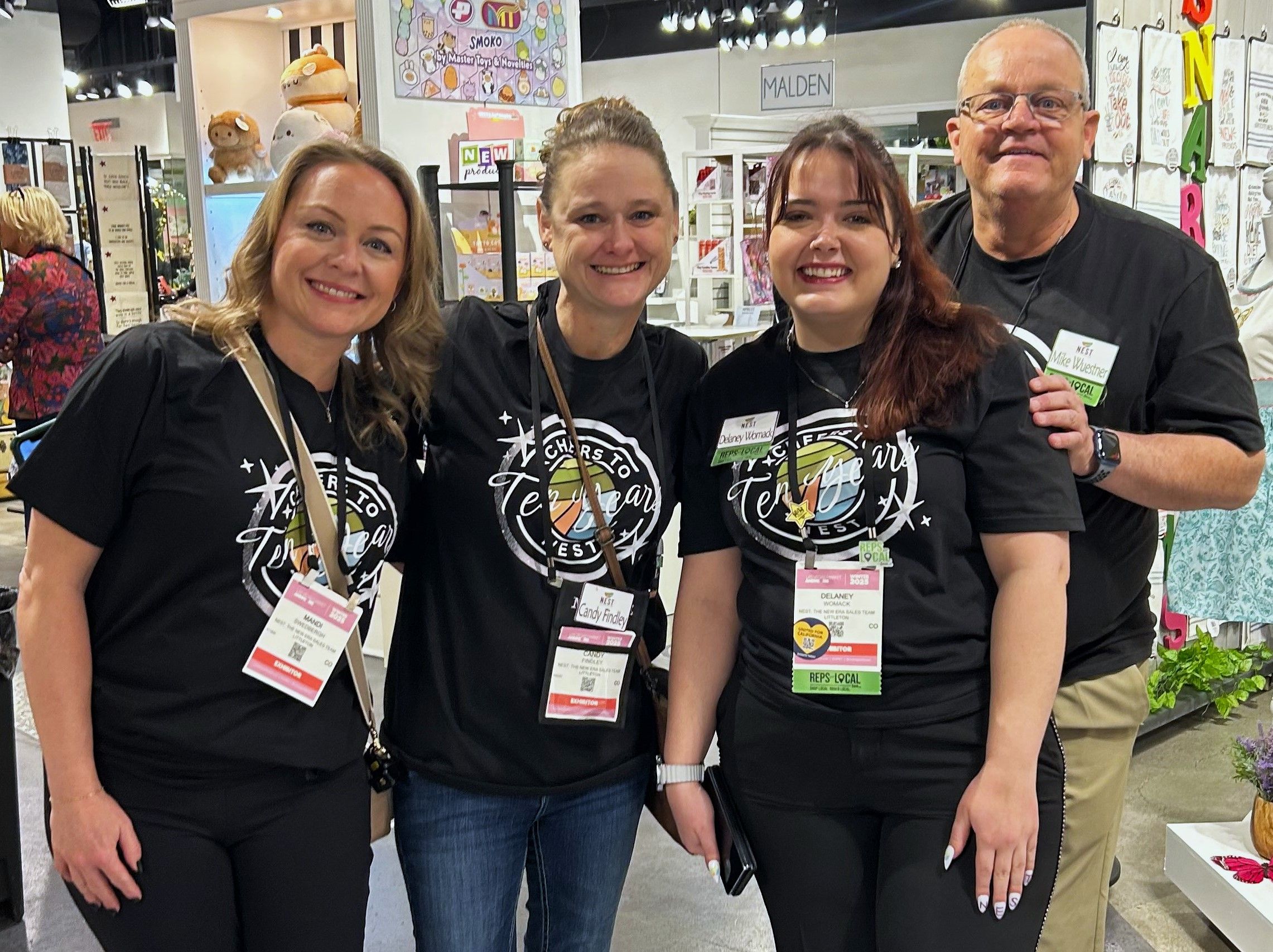 Group photo of three women and one man wearing matching “Cheers to Ten Years” shirts at a trade show. They are smiling in front of a booth featuring gifts and home decor, with lanyards and name badges.