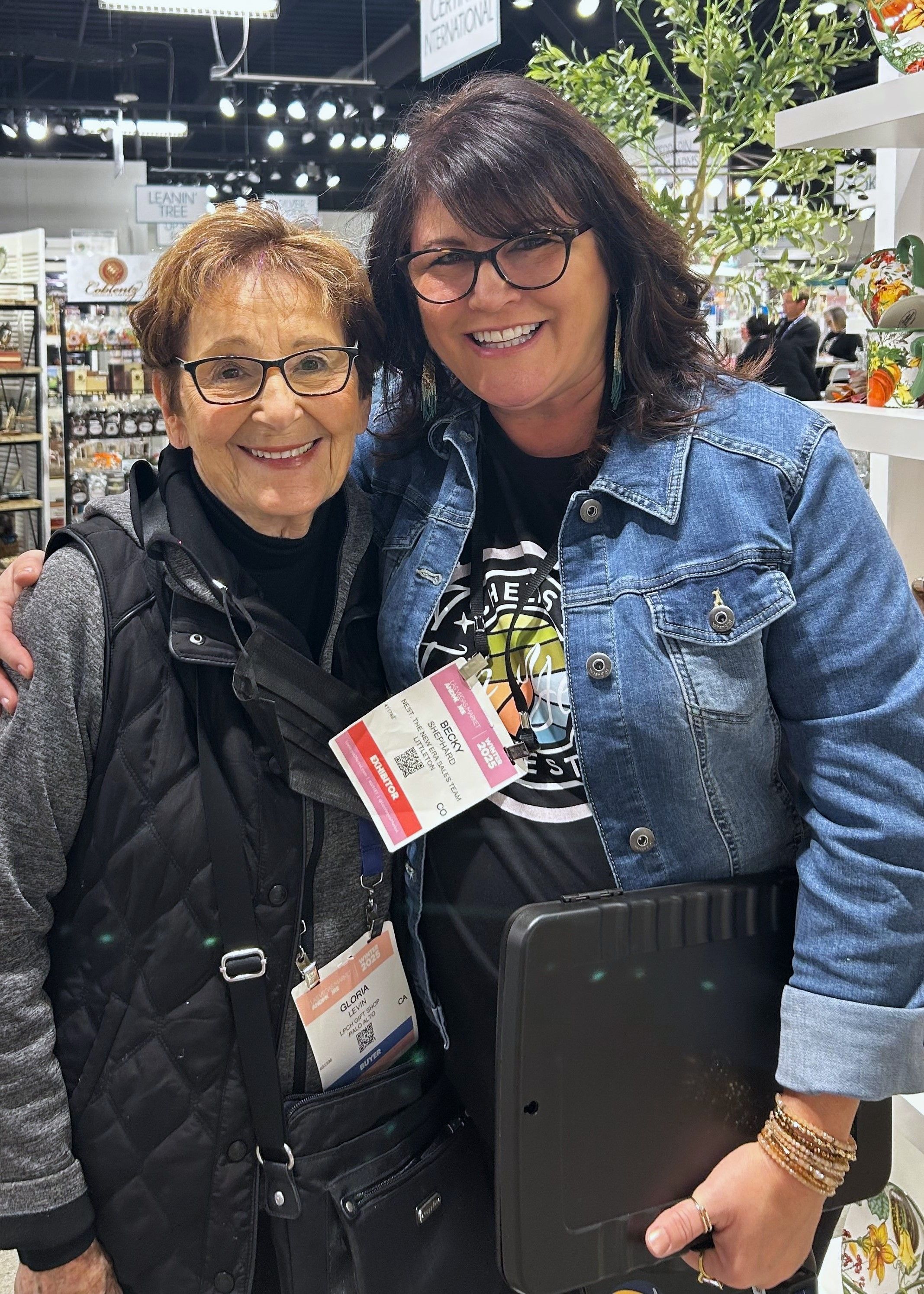 Two women posing in front of retail display shelves; one wearing a denim jacket and show badge, the other in a black vest. They’re surrounded by kitchenware and signage from various brands.