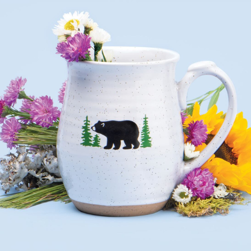 White speckled ceramic mug featuring a black bear and green pine trees illustration, surrounded by a colorful arrangement of wildflowers including daisies, sunflowers, and asters on a soft blue background.