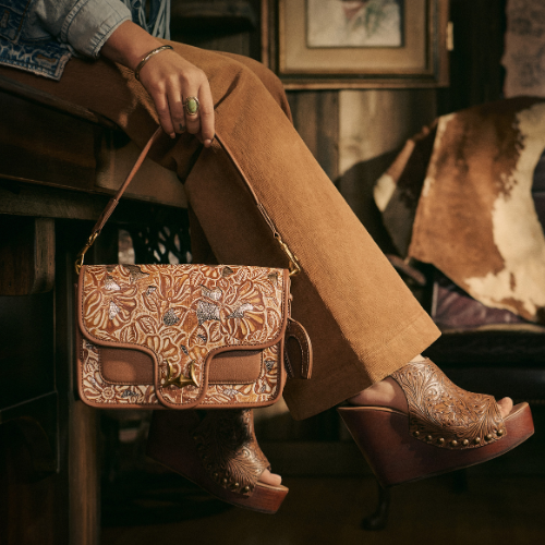 Stylish woman seated indoors wearing brown corduroy pants and tooled leather platform sandals, holding a coordinating embossed leather handbag with intricate floral patterns, set in a rustic, Western-themed room.
