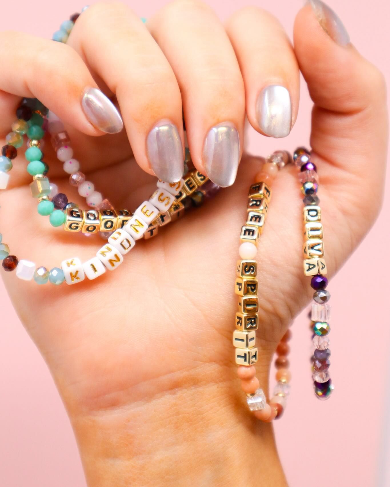 Close-up of a hand with iridescent silver nails holding several colorful beaded bracelets, each featuring inspirational words like “KINDNESS,” “FREE SPIRIT,” “DIVA,” and “GOODNESS.”