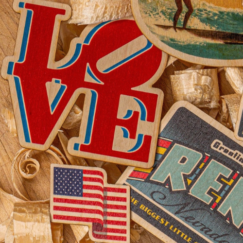 Close-up of layered wooden cutouts featuring a red, white, and blue "LOVE" sign, an American flag, a vintage Reno postcard, and decorative wood shavings in the background.