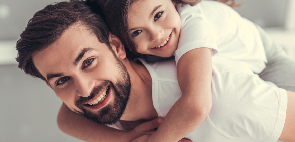 Smiling father giving piggyback ride to his cheerful daughter indoors, both wearing white shirts in a cozy room.