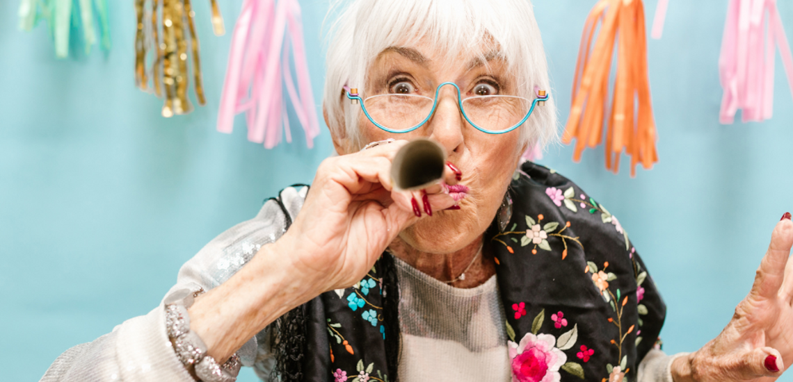 Joyful elderly woman wearing a party hat and blowing a noisemaker in front of colorful tassel garland.